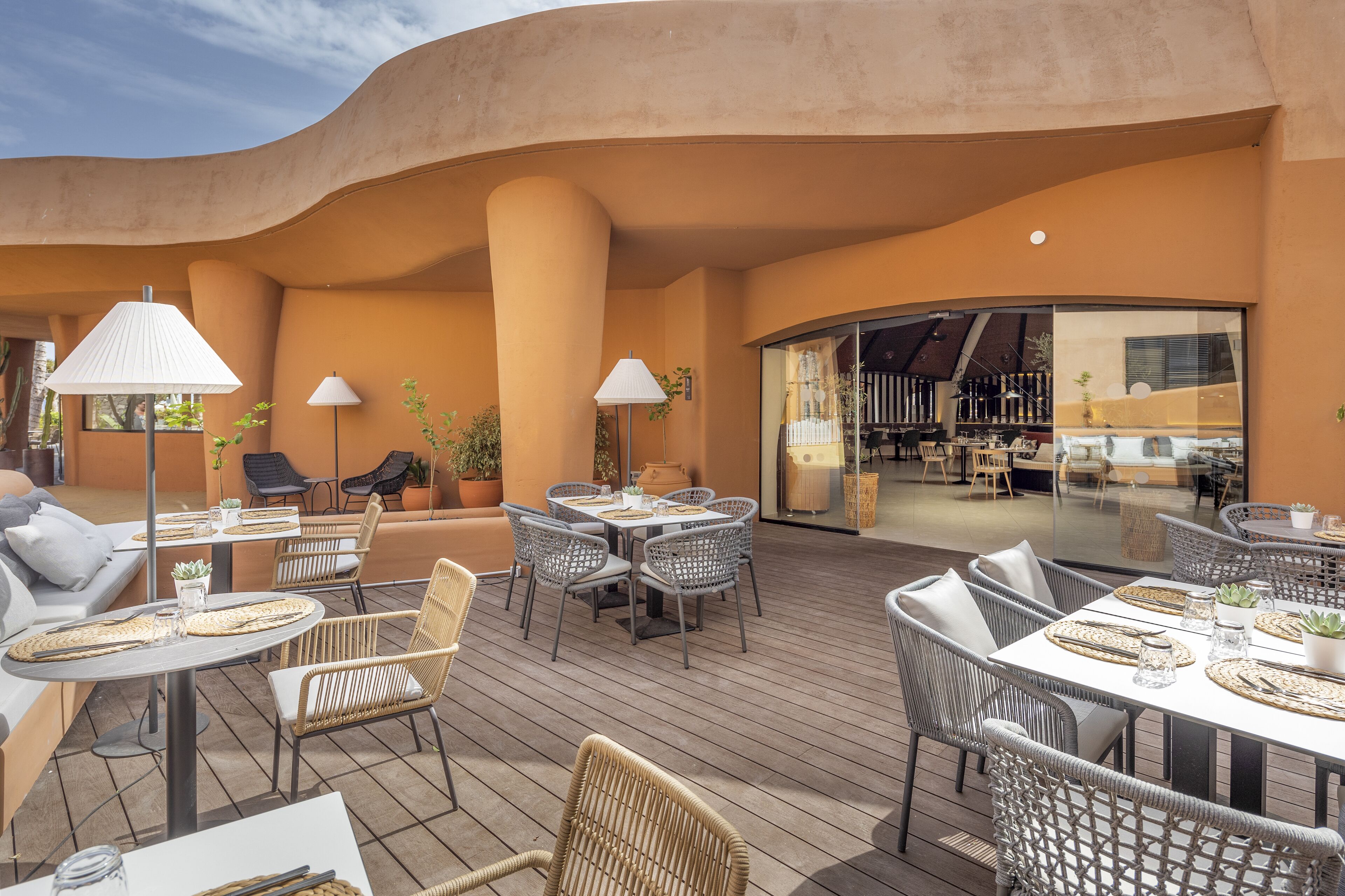 An outdoor patio area with round tables and wicker chairs, a sofa, and a bar area visible through a glass wall.