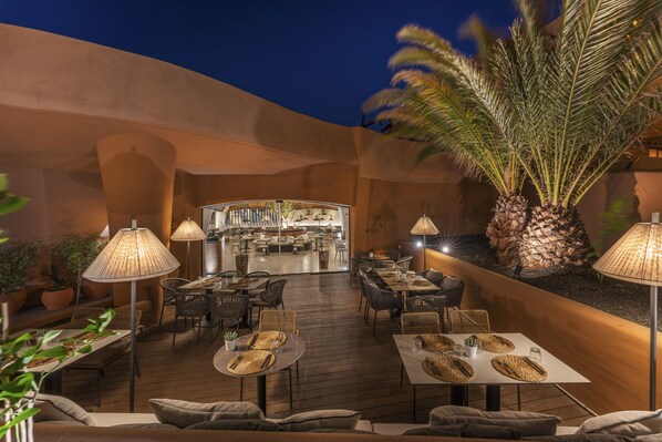 Un espacio exterior para comer con mesas y sillas, un bar al fondo y palmeras.