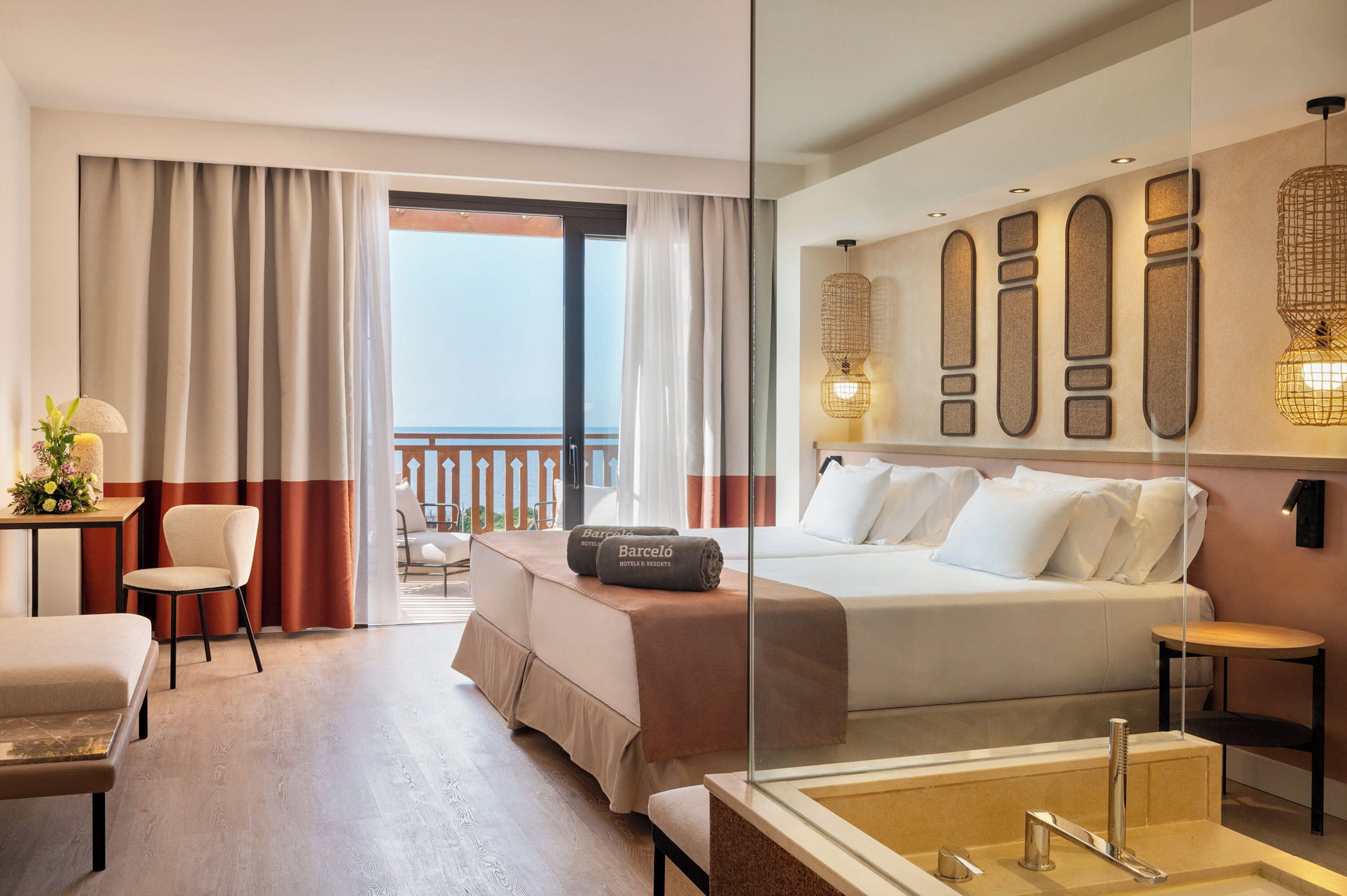 Habitación de hotel con cama, escritorio con lámpara, silla, baño con bañera y balcón con cortinas.
