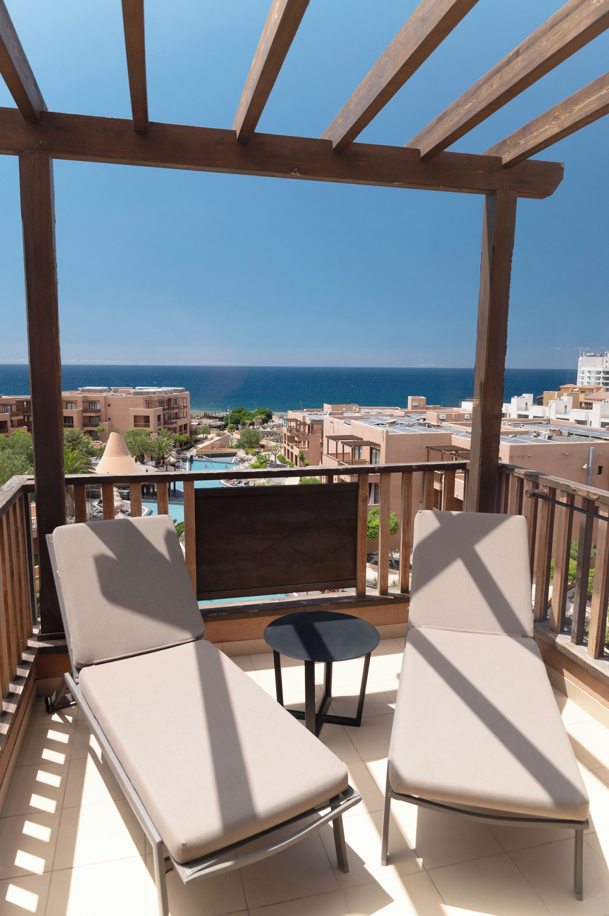 Varanda com duas cadeiras de descanso e uma mesa pequena, com vista para uma piscina e edifícios.