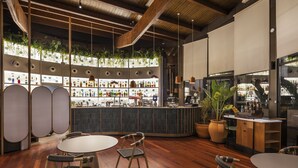 Un espace bar moderne avec un plancher en bois, des tables rondes et des chaises. On y trouve des étagères remplies de bouteilles et une grande plante en pot.