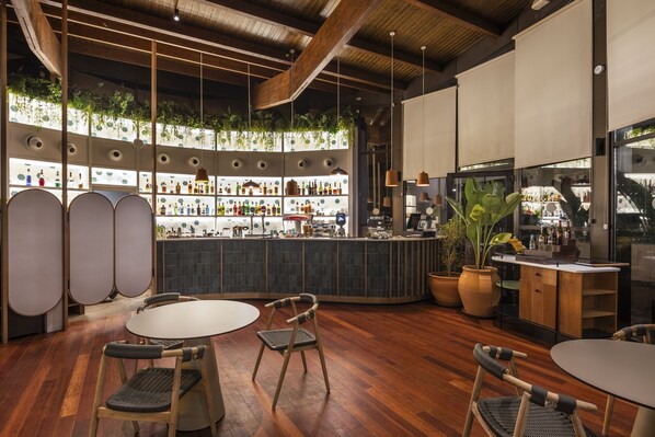 A modern bar area with wooden flooring, round tables, and chairs. There are shelves with bottles and a large potted plant. - Barceló Tenerife Royal Level (San Miguel de Abona)