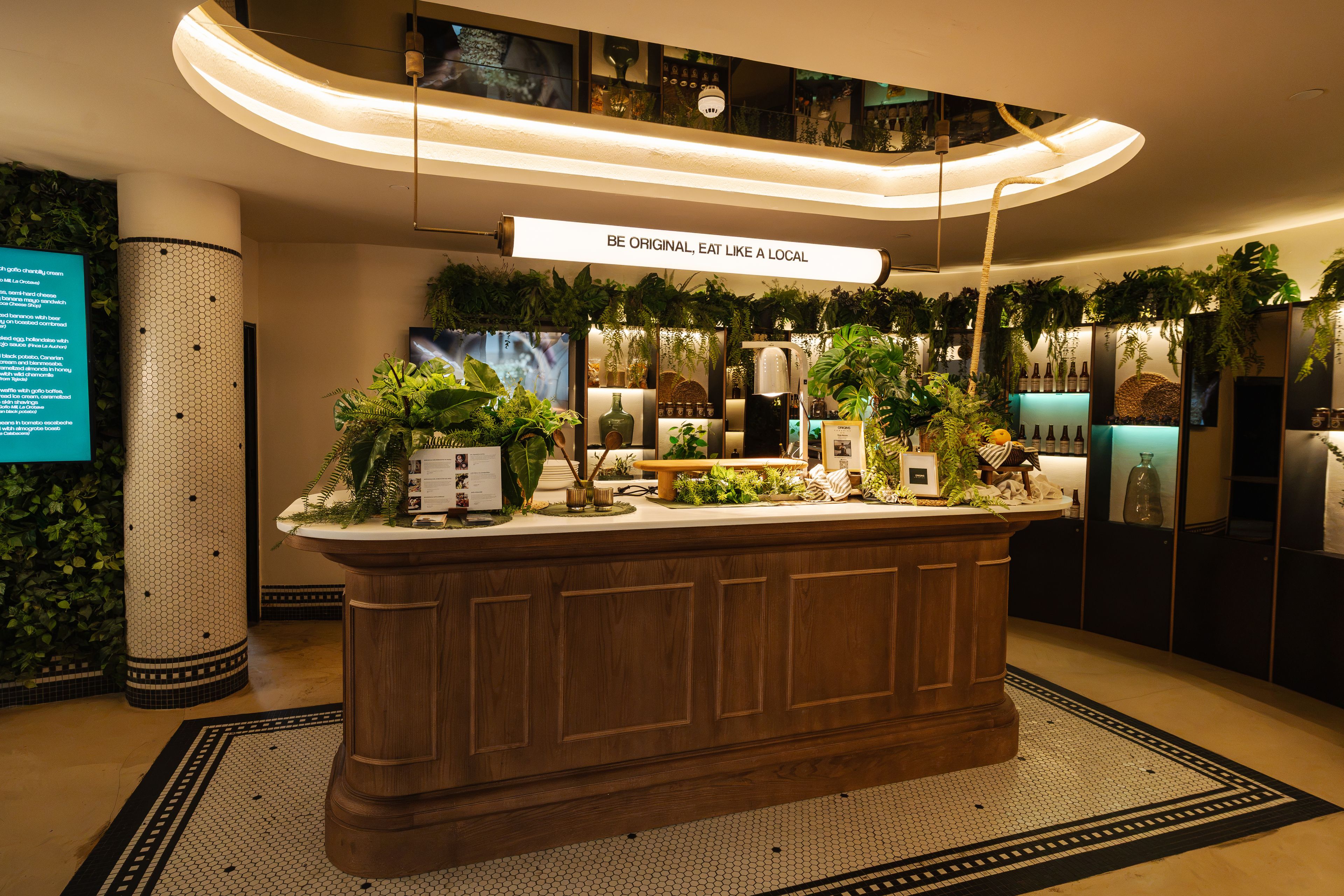 A reception area with a wooden reception desk, a sign above stating 'BE ORIGINAL, EAT LIKE A LOCAL', and a wall decorated with plants and wine bottles.