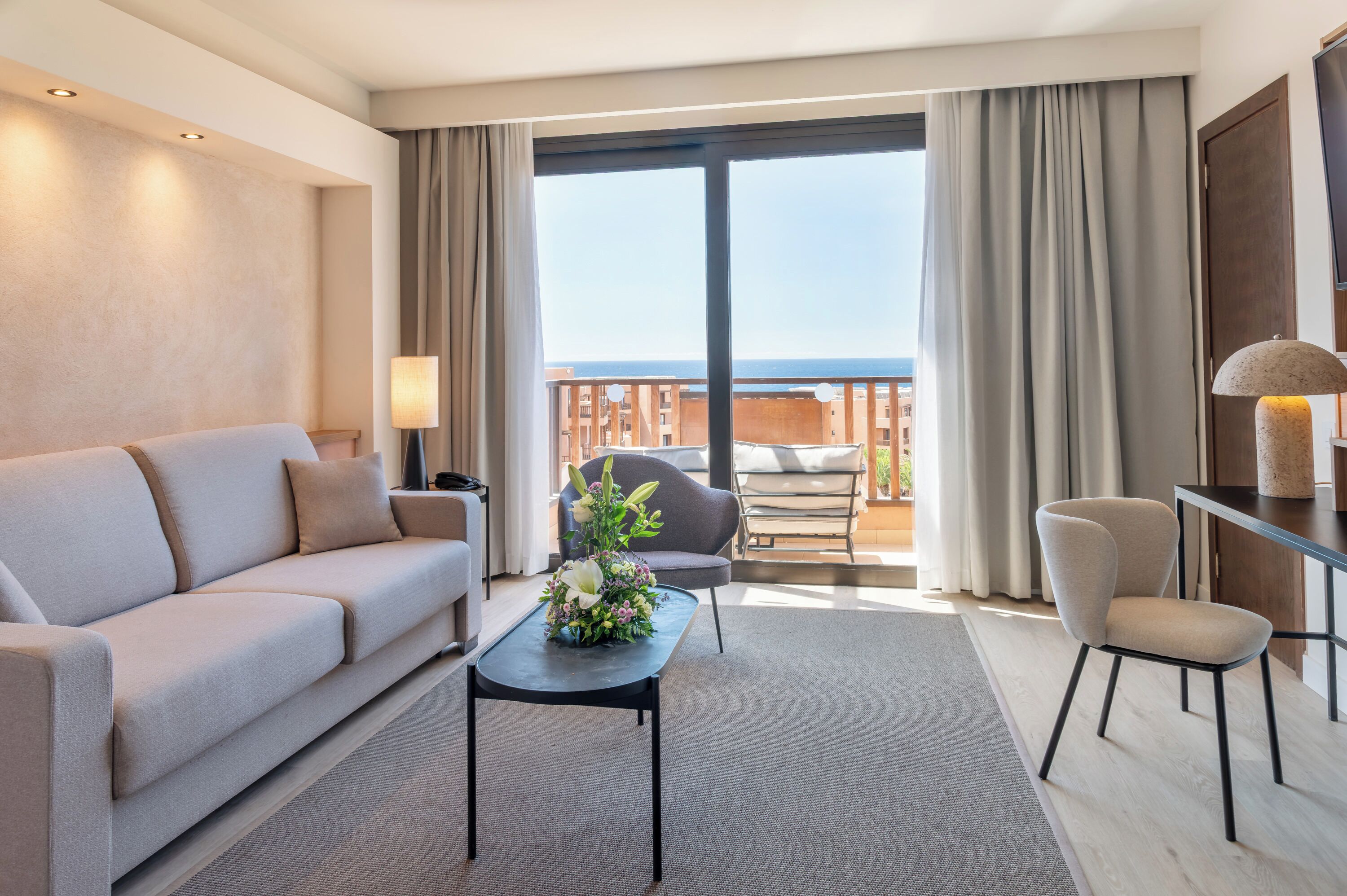 Una habitación de hotel moderna con sofá, una mesa con un arreglo floral, una silla y un balcón con vistas.