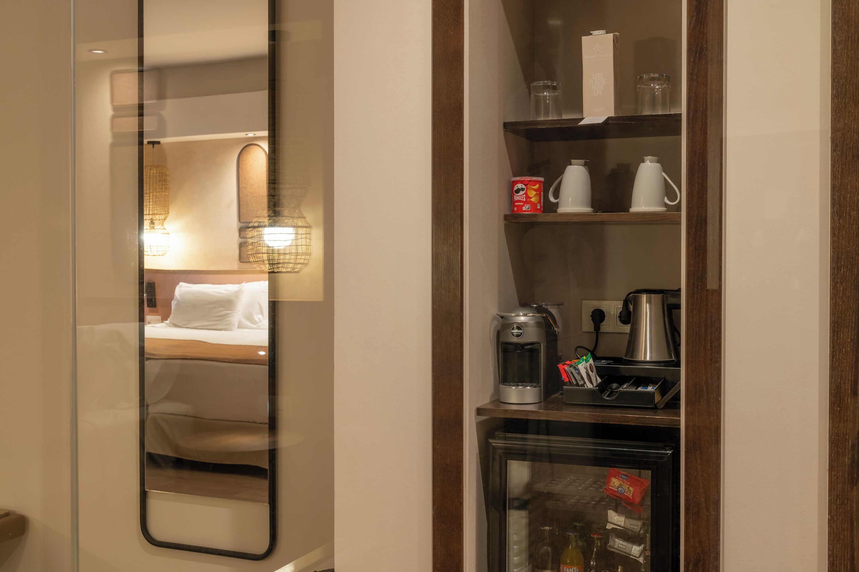 Une chambre d’hôtel avec un lit, un miroir, une étagère avec une machine à café, une bouilloire et un verre d’eau.