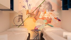 Une chambre d’hôtel avec deux lits, une petite table ronde et une fresque murale représentant un vélo, des fleurs et une lune.