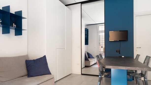 Un salon moderne comprenant un canapé, une table à manger avec des chaises, un téléviseur fixé au mur et une porte-fenêtre coulissante donnant sur une autre pièce.