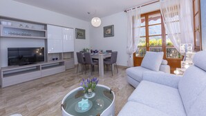 A modern living room with a sofa, a glass coffee table with decorative items, a TV on a built-in shelf, a dining area with a table and chairs, and a large window with curtains. - Carlazzo Sant' Agata Apt. 01 (Carlazzo)