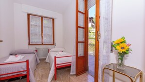 A room with two beds, a window with curtains, a radiator, and a balcony with a glass table and a vase of flowers. - Carlazzo Sant' Agata Apt. 01 (Carlazzo)