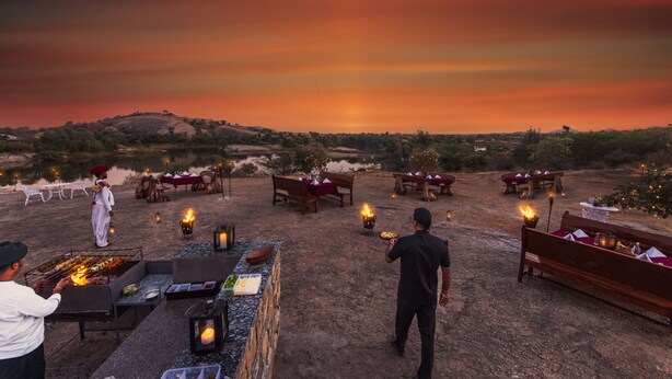 An outdoor dining area with multiple tables and benches, surrounded by a natural landscape and a sunset sky. - WelcomHeritage Cheetagarh Resort and Spa (Bali)