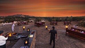 An outdoor dining area with multiple tables and benches, surrounded by a natural landscape and a sunset sky. - Cheetagarh Resort & Spa (Bali)