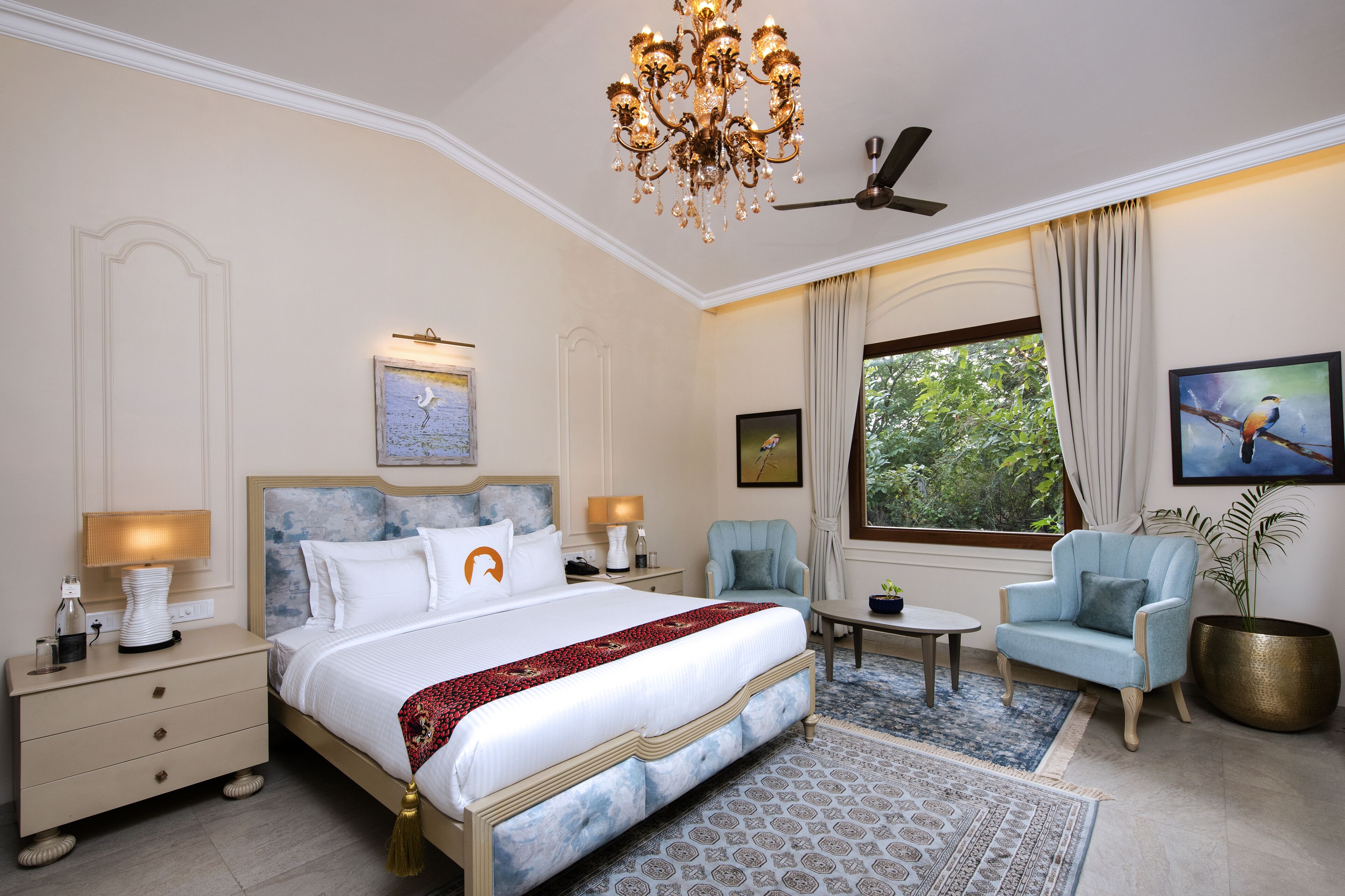 Habitación de hotel con una cama grande, dos mesitas de noche, un candelabro, un ventilador de techo, una ventana con cortinas y vistas a zonas verdes.