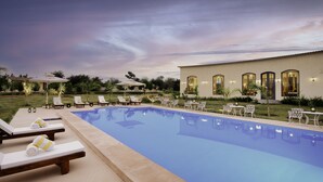 A pool area with lounge chairs and tables, a building with arched windows, and a well-maintained lawn. - WelcomHeritage Cheetagarh Resort and Spa (Bali)