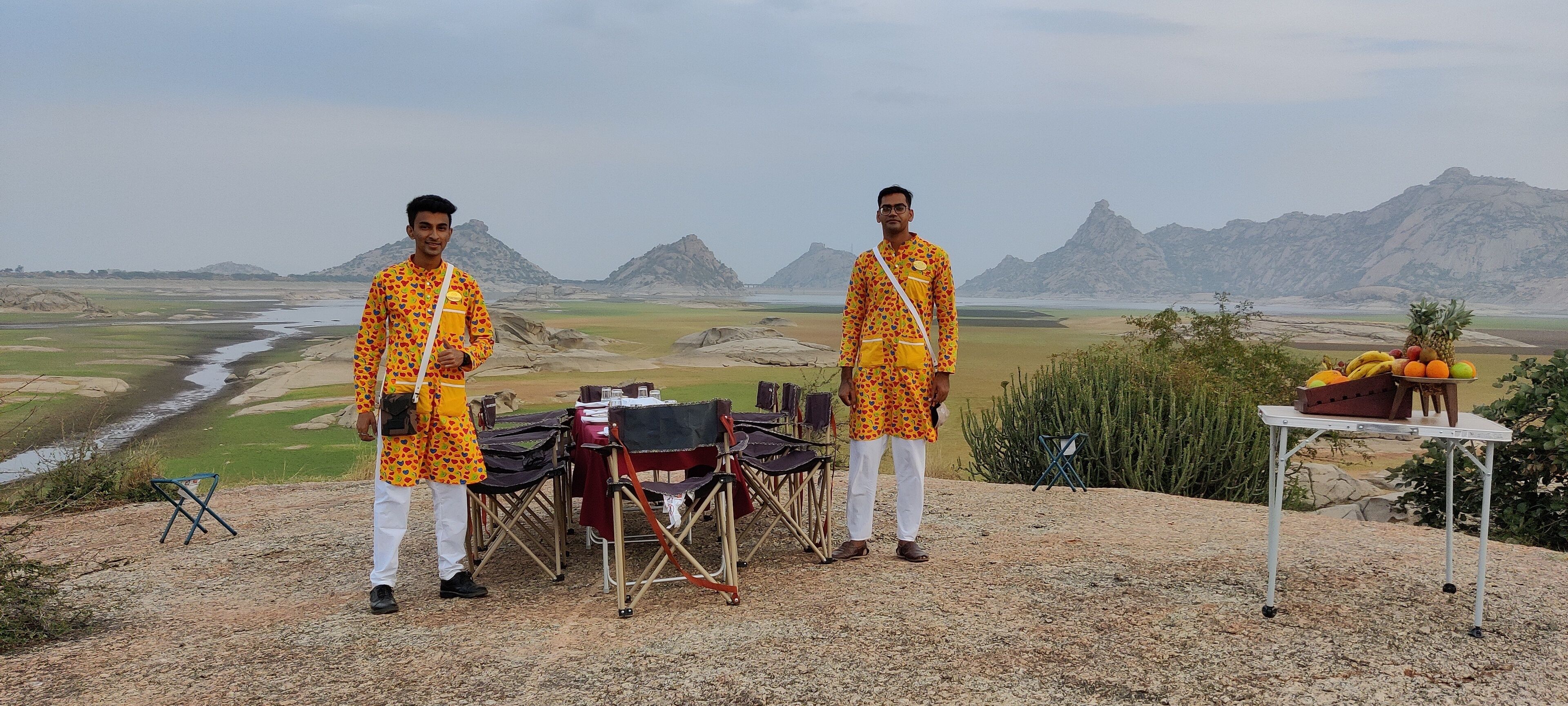 Two individuals standing in front of a table with a view of a river and mountains in the background.