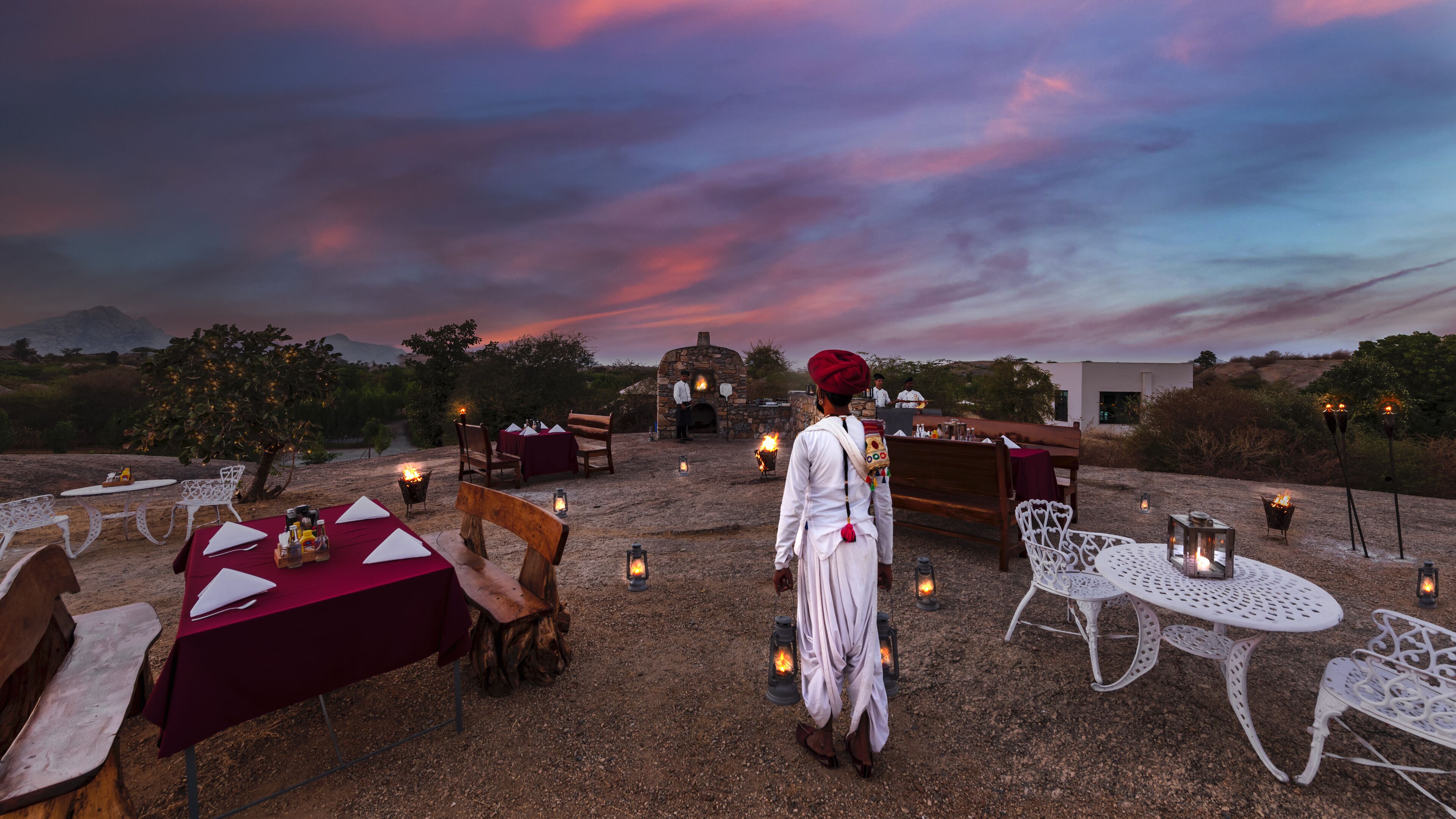 An outdoor dining area with tables and chairs, a stone oven, and a person in traditional attire.