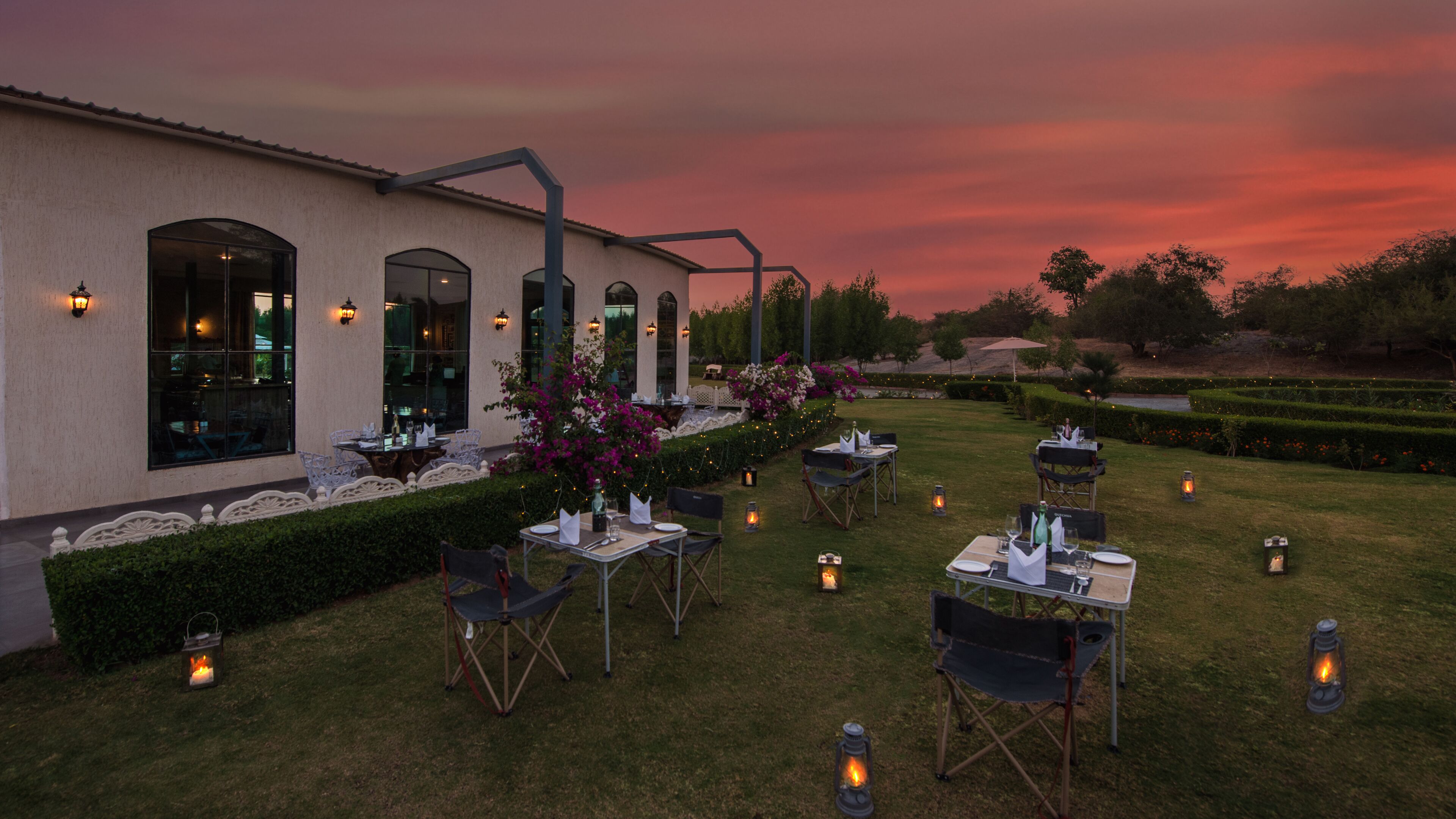 An outdoor dining area with tables and chairs arranged on a lawn, surrounded by a building with arched windows and a garden with flowering plants.