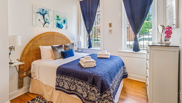 Ein ordentlich eingerichtetes Schlafzimmer mit einem großen Bett, einem Nachttisch mit Lampe, einer Kommode und Bildern an der Wand.