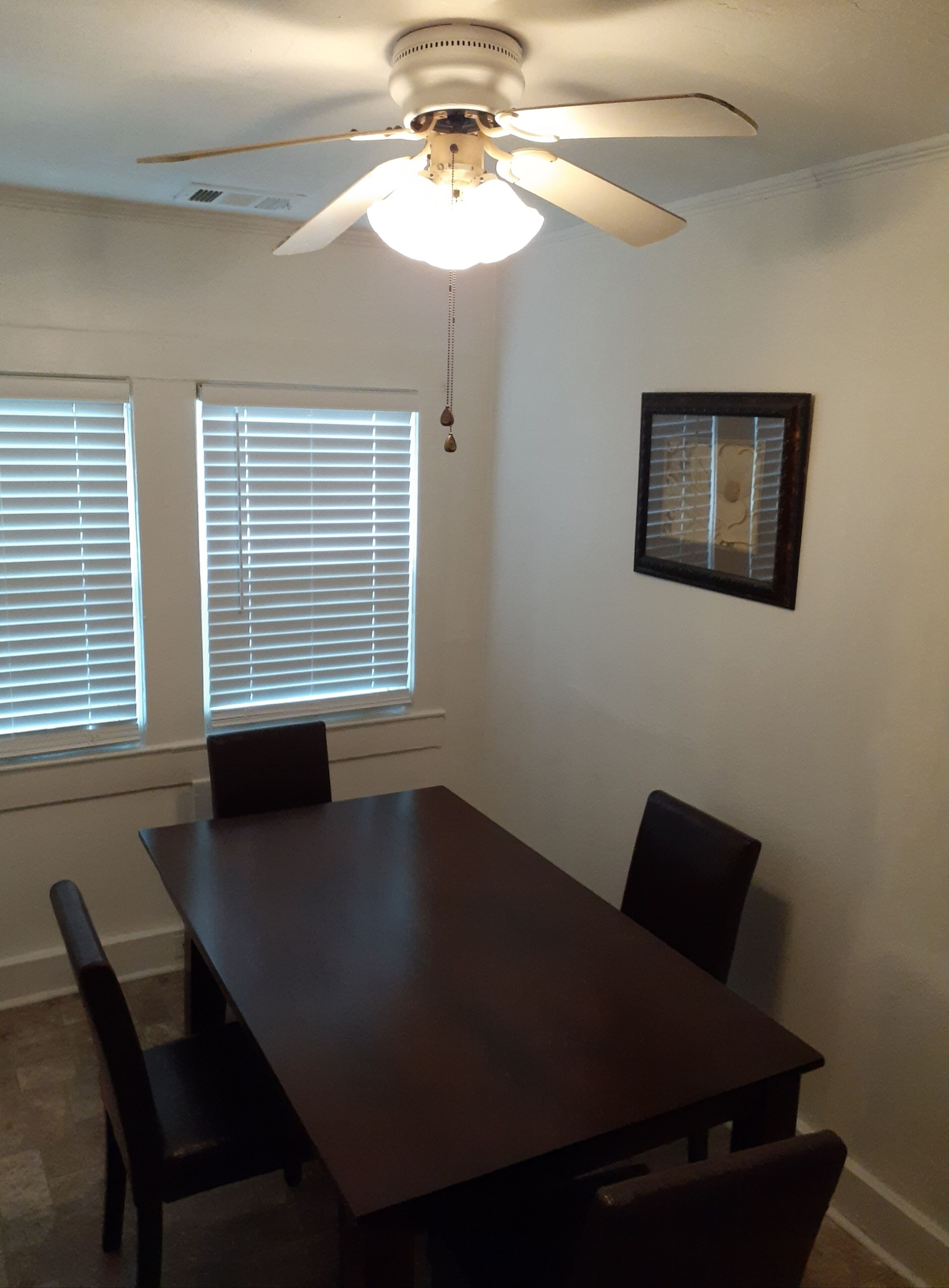 A dining area with a dark wooden table and chairs, a ceiling fan, and two windows with blinds.