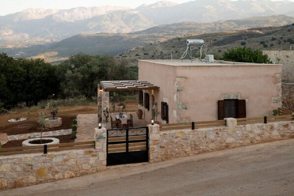 A small building with a stone wall and a gate, surrounded by greenery and mountains in the background.