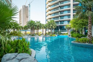 A swimming pool surrounded by palm trees and greenery, with modern buildings in the background.