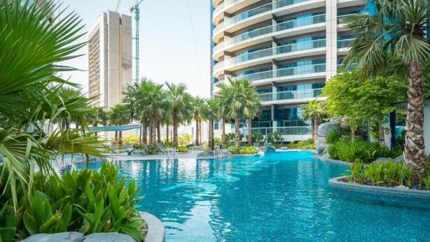 A swimming pool surrounded by palm trees and greenery, with modern buildings in the background. - Cool Dubai Apt next Burj Khalifa & Design District (Dubai)