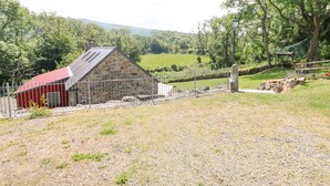 A stone building with a red roof, surrounded by a gravel area and a metal fence. - Trewrach Cottage (Newport)