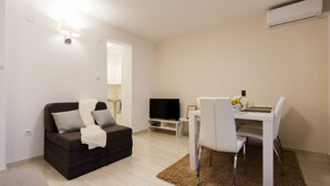 Ein modernes Wohnzimmer mit einem dunklen Sofa, einem weißen Esstisch mit Stühlen, einem Flachbildfernseher und einer kleinen TV-Konsole.