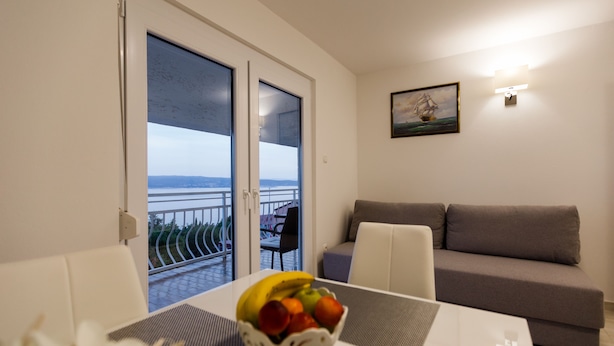 Ein modernes Wohnzimmer mit grauem Sofa, einem Esstisch mit Obstschale und einem Balkon mit Meerblick.