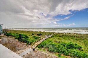 Blick auf die Küste mit einer Holzpromenade, die zu einem Strand führt, einem Rettungsschwimmturm und einer Rasenfläche mit Büschen.