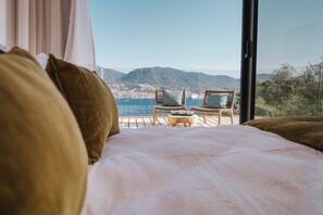 A bedroom with a large bed, two wicker chairs, and a view of the sea and mountains.