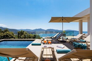 A poolside area with lounge chairs, a table with drinks, and a view of mountains and a beach.