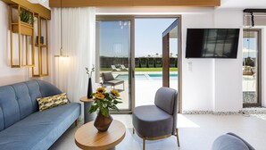 Een moderne woonkamer met een bank, fauteuil en een ronde houten tafel met een vaas zonnebloemen. Een grote televisie is aan de muur bevestigd en er is een schuifdeur naar een buitenruimte met een zwembad en lounge stoelen.