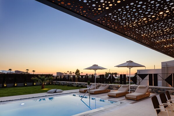 A pool area with lounge chairs and umbrellas, surrounded by a landscaped garden and a view of the sunset.