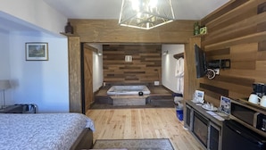 A bedroom with a bed, a wooden floor, a small bathroom, a kitchenette, and a shower area. - Country Suite - Private HotTub, KingBd, Downtown (Wolfville)