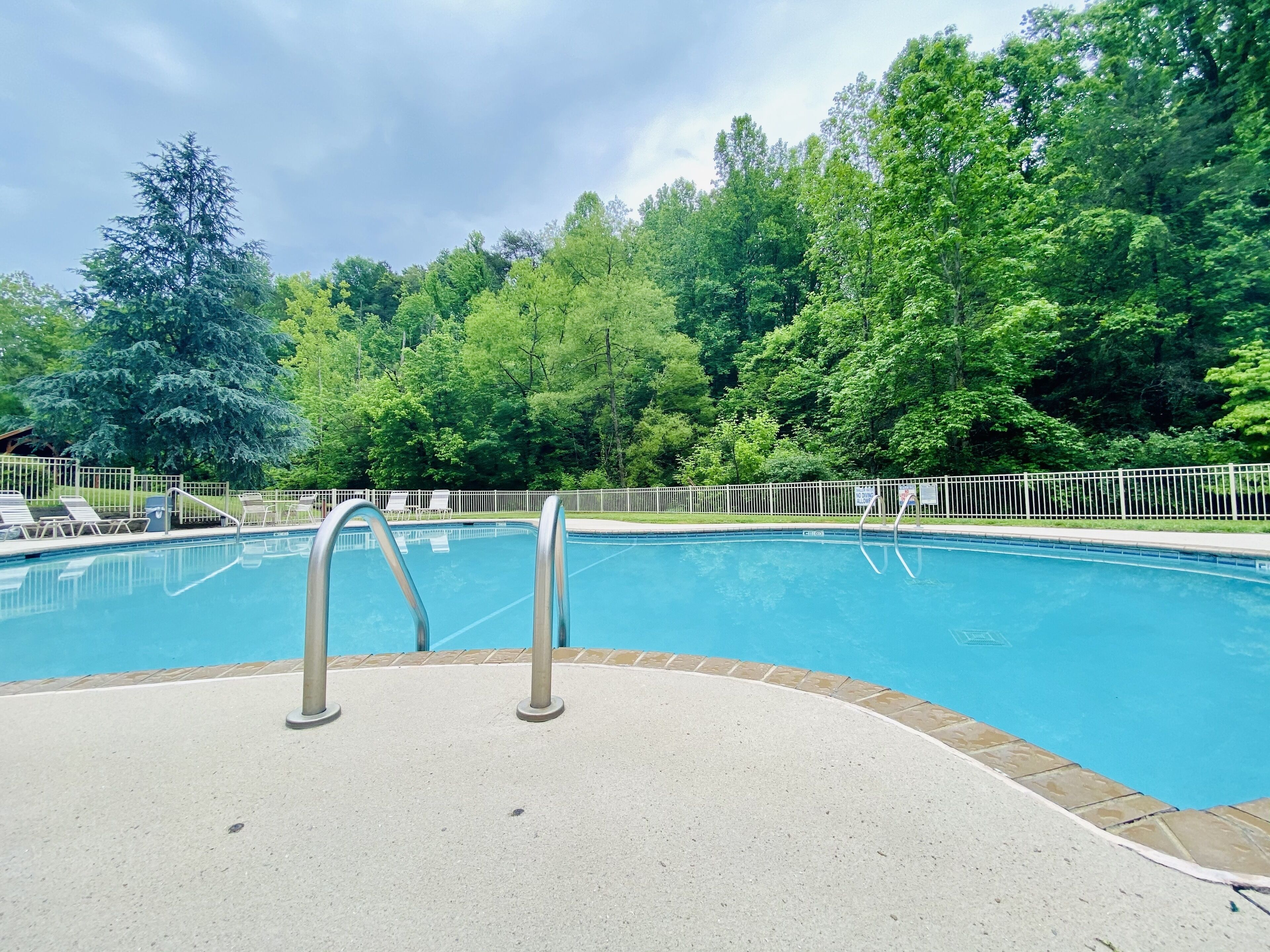 A swimming pool with metal ladders, surrounded by a white fence and lush green trees.