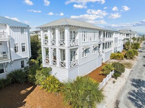 A multi-story white building with balconies, surrounded by greenery and a clear sky.