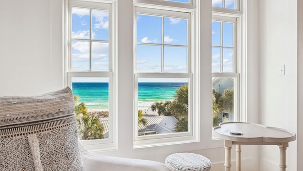 Ein Zimmer mit großem Fenster und Blick auf einen Strand, einem weißen Tisch und einem gemusterten Kissen.