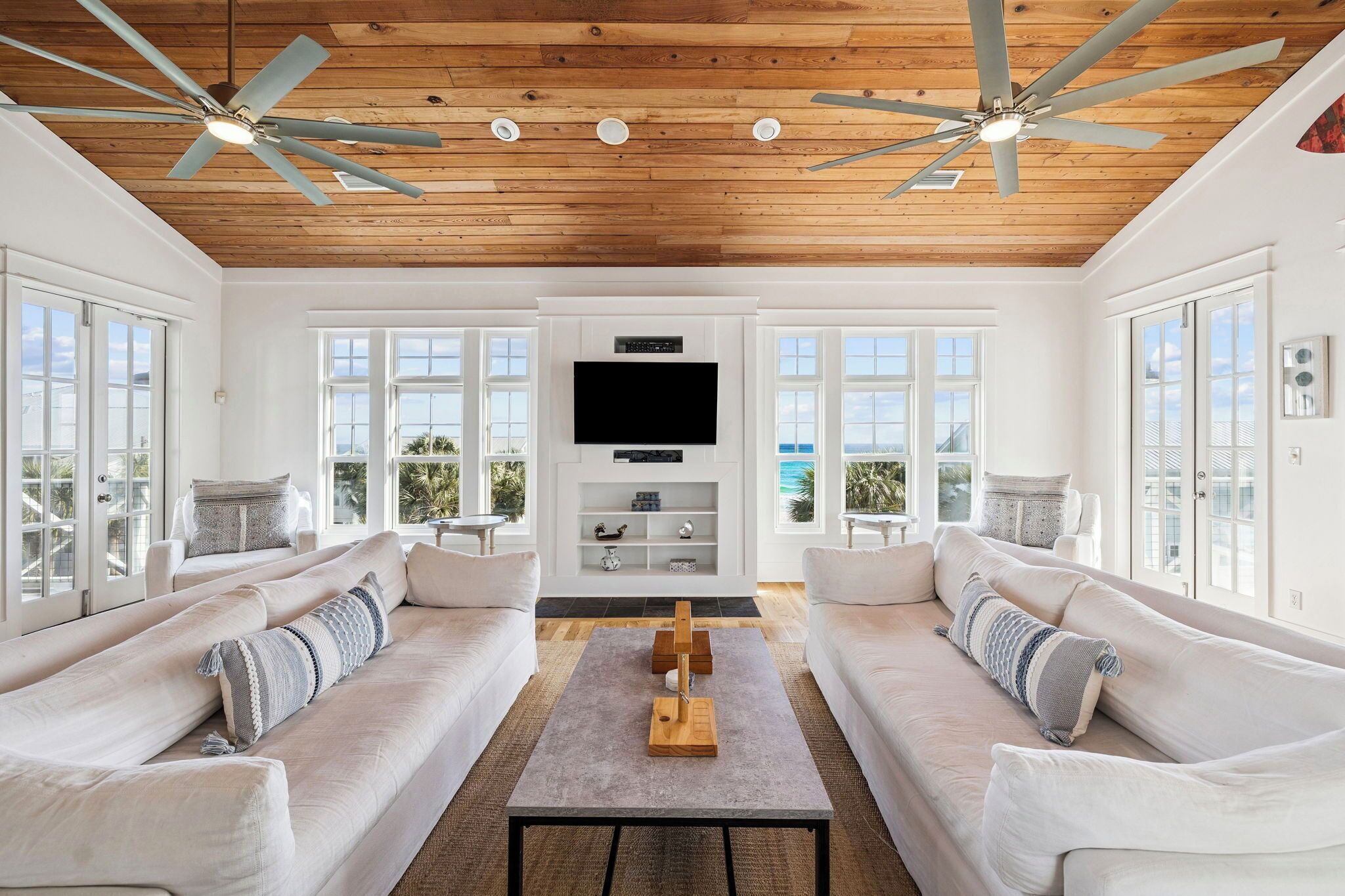 A spacious living room with two white sofas, a central coffee table, a flat-screen TV, ceiling fans, and large windows offering a view.