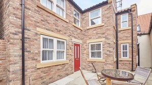 Une maison en briques avec une porte rouge, deux fenêtres à cadres blancs et une table ronde entourée de chaises dans la cour.