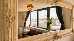 Una habitación con un espejo grande y ornamentado, un sofá de cuero marrón, un jarrón con flores y vistas a un paisaje urbano a través de una ventana con cortinas negras.