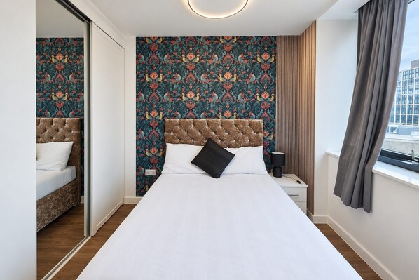 A hotel room with a bed, a nightstand, a mirror, and a window with curtains. - 2 Bedroom Central Apartment  (Liverpool)