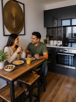 Una cocina moderna con muebles oscuros, un horno de acero inoxidable y una mesa de comedor con dos personas comiendo.