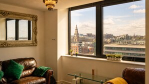 A room with a leather sofa, a glass table, a large window with a city view, and a chandelier.