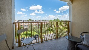 A balcony with a view of a parking lot and buildings, a chair, a table, and a railing.