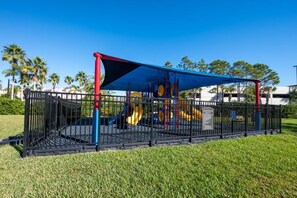 Ein Spielplatz mit blauem Baldachin und einem umzäunten Bereich, umgeben von Gras und Palmen.