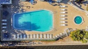 Vista aèria d'un complex turístic amb una gran piscina rectangular, una piscina circular més petita i diverses zones de relaxació amb gandules.