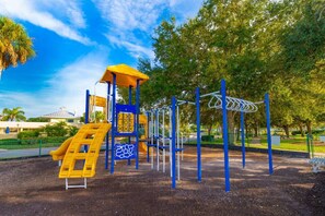 Kids can play on the slide, climbing elements, and netted climbing frame in this open playground area surrounded by trees.