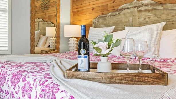Ein Schlafzimmer mit Holz-Kopfteil, einem Bett mit floralem Bettzeug, einem Nachttisch mit Lampe und einem Tablett mit Flasche und Gläsern.