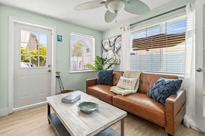 Un salon avec un canapé en cuir marron, une table basse en bois, un ventilateur de plafond et une fenêtre avec des stores.