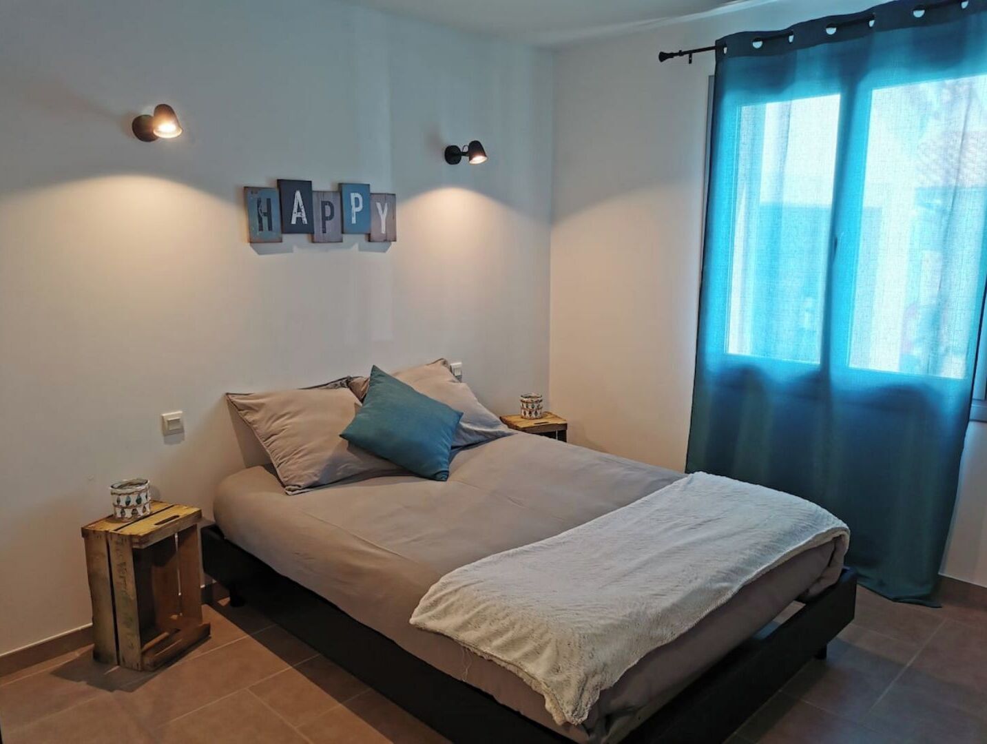 Une chambre à coucher avec un lit, des oreillers, une table de chevet, une lampe fixée au mur et un rideau.